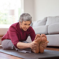Beautiful senior woman doing stretching exercise while sitting on yoga mat at home. Mature woman exercising in sportswear by stretching forward to touch toes. Healthy active lady doing yoga.